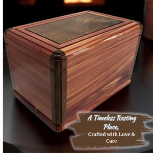 Op de afbeelding: Een houten doos met een donkerhouten deksel en een licht houten lichaam. De doos heeft een donkerhouten rand rond de randen. De doos staat op een donker oppervlak. De tekst "A Timeless Resting Place, Crafted with Love & Care" staat op een bruine achtergrond in de rechterbenedenhoek van de afbeelding.