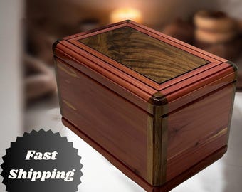 May include: A rectangular wooden box with a dark wood top and a lighter wood base. The box has a decorative border around the top and bottom.