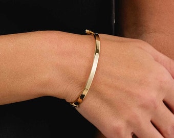 May include: A gold-colored bangle bracelet with a simple, elegant design. The bracelet is a solid band with a clasp closure. The smooth, polished surface reflects light, creating a bright, shiny appearance. The bracelet is shown on a person's wrist.