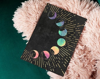 May include: A black card with a gold design of a crescent moon with a rainbow swirl pattern. The card is on a pink fuzzy surface.