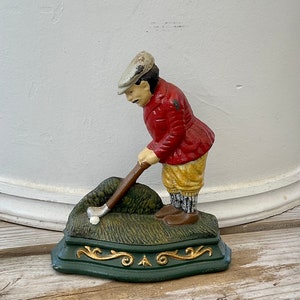 May include: A cast iron doorstop shaped like a golfer in a red jacket, white hat, and yellow pants. The golfer is swinging a club and a white golf ball is on the green grass base.