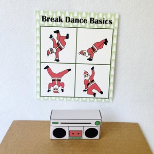 May include: A white paper boombox sits on a brown surface below a card with the text "Break Dance Basics." The card features four illustrations of Santa Claus breakdancing in red suits and white beards. The card has a green border with white Christmas trees.