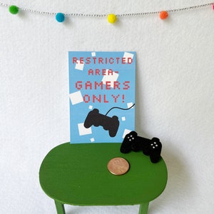 May include: A green miniature table holds a sign that reads "RESTRICTED AREA - GAMERS ONLY!" in red pixelated text. A black controller graphic is on the sign. A black controller and a penny are on the table. A string of colorful pom-poms hangs above.