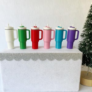 May include: Six colorful travel mugs with handles and straws are arranged on a white surface. The mugs are white, green, red, pink, teal, and purple. Each mug has a white lid and a handle. A small Christmas tree is in the background.