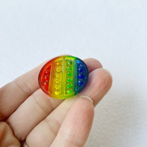 May include: A rainbow-colored, oval-shaped fidget toy with a bubble-popping design. The toy features a spectrum of colors, including red, orange, yellow, green, and blue. The surface has a series of small, raised bubbles.