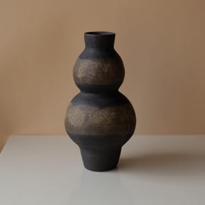 Könnte beinhalten: Ein Set aus drei Keramikbechern mit gesprenkelter, matter Oberfläche. Die Becher sind in Creme-, Hellgrau- und Dunkelgrautönen gehalten. Jeder Becher hat ein schlichtes, minimalistisches Design.