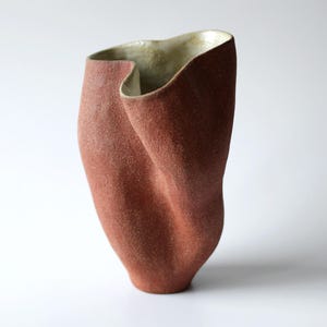 Może przedstawiać: Wysoki, ręcznie formowany wazon ceramiczny o teksturowanej powierzchni w kolorze terakoty i gładkim, jasnokremowym wnętrzu. Naczynie posiada nieregularną, falistą krawędź oraz pofałdowaną, organiczną sylwetkę, która zwęża się ku podstawie.