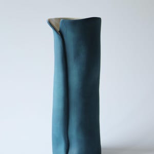 Può includere: Un vaso cilindrico alto in ceramica con una finitura esterna opaca color blu ottanio profondo. Il vaso presenta una distinta piega verticale su un lato, creando una silhouette organica unica con un bordo superiore aperto e leggermente asimmetrico.