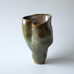 Può includere: Un vaso in ceramica realizzato a mano con una forma organica irregolare e una finitura lucida. L'esterno presenta toni sfumati di verde oliva, marrone e accenni di blu, mentre l'interno rivela una superficie smaltata liscia color malva.