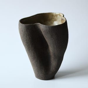 Può includere: Un vaso in ceramica fatto a mano con un esterno marrone scuro testurizzato e un interno liscio color beige chiaro. La forma è asimmetrica e organica, caratterizzata da un bordo pizzicato e ondulato che crea una silhouette irregolare.