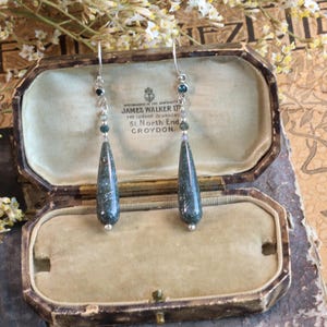 May include: A pair of drop earrings featuring elongated, dark green, speckled stone pendants suspended from silver-toned hooks. Each earring includes a small chain with tiny, dark blue and clear beads connecting the hook to the main stone drop.
