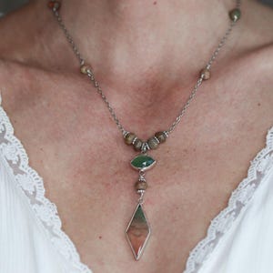 May include: A silver chain necklace with brown and green gemstone beads. The necklace has a long pendant with a green gemstone at the top and a large, diamond-shaped, multi-colored gemstone at the bottom.
