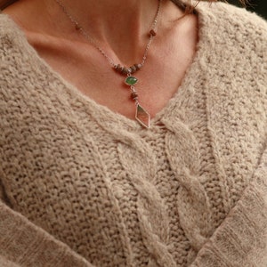 May include: A beige cable knit sweater with a silver necklace featuring green and brown stones.
