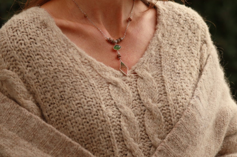 May include: A beige cable knit sweater with a silver necklace featuring green and brown stones.