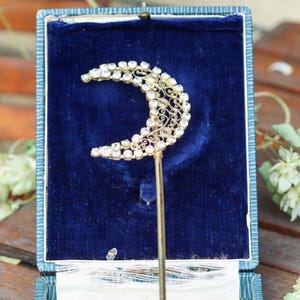 Pode incluir: Um gancho de cabelo dourado com um design de meia-lua adornado com strass transparentes e brilhantes e filigrana metálica intrincada. A meia-lua está presa a uma haste metálica longa e fina, apresentada dentro de uma caixa forrada a veludo azul.