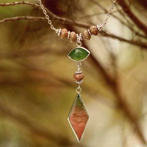 May include: A silver necklace with a green and pink gemstone pendant. The pendant is shaped like a teardrop and is surrounded by smaller brown beads. The necklace is hanging from a branch.
