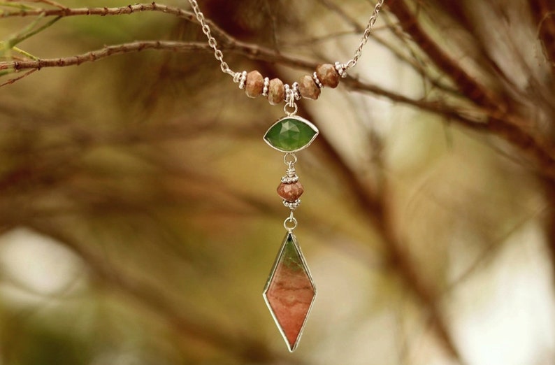 May include: A silver necklace with a green and pink gemstone pendant. The pendant is shaped like a teardrop and is surrounded by smaller brown beads. The necklace is hanging from a branch.
