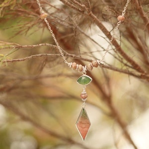 May include: A silver chain necklace with brown and green gemstone beads and a pendant featuring a green and pink gemstone. The necklace is hanging from a branch.