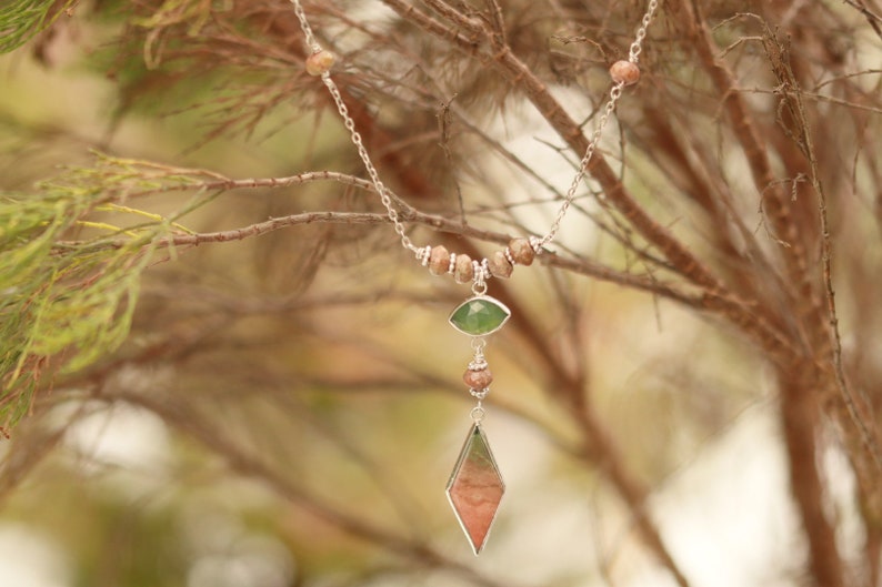 May include: A silver chain necklace with brown and green gemstone beads and a pendant featuring a green and pink gemstone. The necklace is hanging from a branch.
