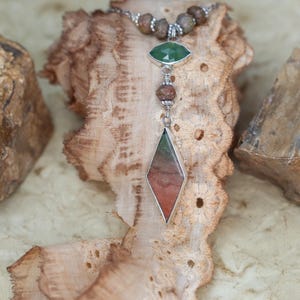 May include: A silver necklace with a brown, green, and red gemstone pendant. The pendant is shaped like a diamond and is surrounded by smaller brown beads. The necklace is displayed on a brown lace fabric.