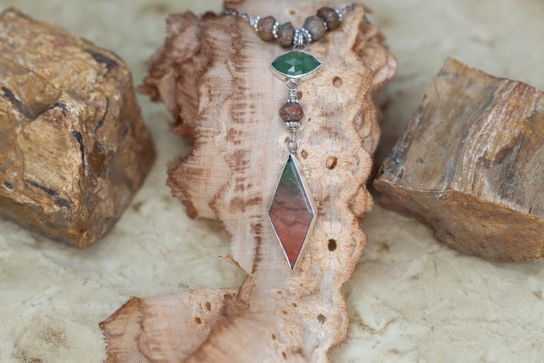 May include: A silver necklace with a brown, green, and red gemstone pendant. The pendant is shaped like a diamond and is surrounded by smaller brown beads. The necklace is displayed on a brown lace fabric.