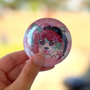 Könnte beinhalten: Ein runder Ansteckbutton mit einer holografischen Sternenmuster-Beschichtung. Die Grafik zeigt eine stilisierte anthropomorphe Figur mit rotem und blaugrünem Fell, großen dunklen Augen und einem lächelnden Ausdruck vor einem rosa-weiß gestreiften Hintergrund.
