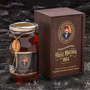 May include: A glass jar of organic chestnut flower honey with a gold lid and a black label. The label has the text "YÖRESEL" and "ORGANIK KESTANE CICEK BALI" in gold lettering. The jar is sitting on a dark marble surface next to a wooden box with a gold label that reads "HAFIZ MUSTAFA 1864 ISTANBUL".