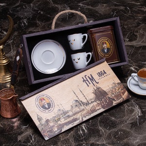 Può includere: Una scatola di legno con un coperchio a cerniera, contenente due tazze da caffè e piattini in porcellana bianca, una lattina di caffè e una targa in legno decorativa con il testo "HM 1864 ISTANBUL".