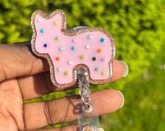 May include: A pink, cat-shaped badge reel with colorful dot accents and a glittery gold border. The retractable reel has a clear plastic strap and a gold glitter button. The background is a blurred green.