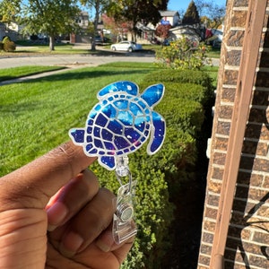 May include: A blue and white turtle-shaped badge reel with a galaxy design. The badge reel has a clear plastic clip and a retractable cord. The turtle design is filled with a starry night sky pattern.