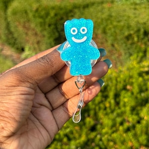Peut inclure: Un enrouleur de badge bleu clair scintillant avec un personnage de dessin animé souriant. L'enrouleur de badge a un cordon rétractable en plastique transparent et un clip en métal. Le fond est un feuillage vert flou.