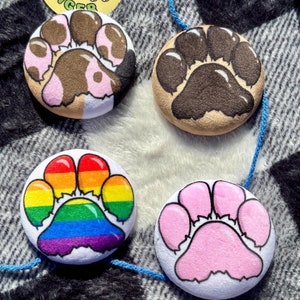 Pode incluir: Uma coleção de botões redondos com pegadas coloridas e cordas azuis. Um botão apresenta uma pegada arco-íris, enquanto outros exibem designs de pegadas marrons, rosa e bege. Um botão amarelo com um tigre e o texto "FRUITY TIGER" também está presente.