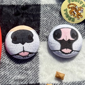 Pode incluir: Dois botões redondos de tecido com desenhos de focinhos de cão, um com a boca fechada e outro com a boca aberta. Um botão amarelo com um gráfico de tigre e as palavras "FRUIT & TIGER" também é visível. Os itens estão em uma superfície xadrez em preto e branco.