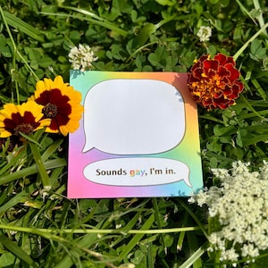 May include: A square, rainbow-edged notepad with a speech bubble shape and the text "Sounds gay, I'm in." is surrounded by green grass and colorful flowers. The notepad is white with a blank space for writing.