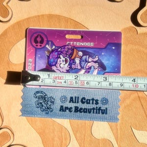 May include: A purple and pink attendee badge with a cartoon cat illustration, the word "ATTENDEE", and the year "2023". A blue ribbon with the text "All Cats Are Beautiful" is below the badge. A measuring tape shows the badge is approximately 4 inches long.