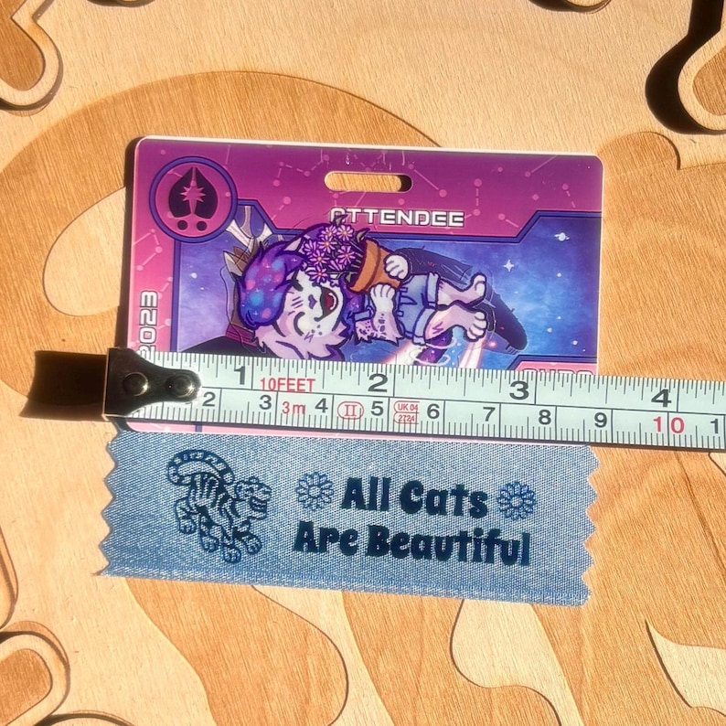 May include: A purple and pink attendee badge with a cartoon cat illustration, the word "ATTENDEE", and the year "2023". A blue ribbon with the text "All Cats Are Beautiful" is below the badge. A measuring tape shows the badge is approximately 4 inches long.
