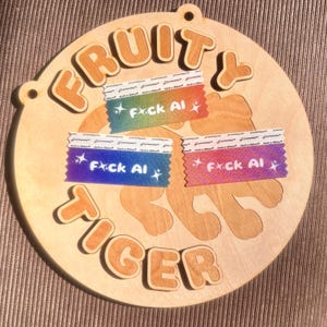 May include: A round wooden sign with the words "FRUITY TIGER" in raised, orange-brown letters. The sign features a tiger shape and three colorful tape strips with the text "F*ck AI". The sign has a natural wood grain.
