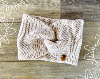 May include: A beige knitted headband with a knotted design and a small brown leather tag.