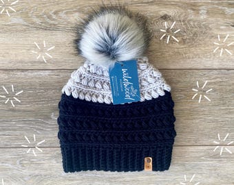 Może przedstawiać: Ręcznie robiona zimowa czapka w kolorze granatowym i szarym z pomponem z imitacji futra. Czapka posiada metkę z napisem "wildwood headwear". Modny dodatek na zimę.