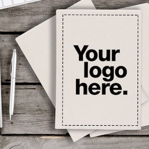 Puede incluir: Un cuaderno blanco con una portada en blanco que dice "Your logo here." El cuaderno está sobre una mesa de madera con un teclado, una planta en maceta, una taza de café, un bolígrafo y un cuaderno espiral.