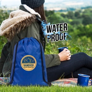 Op de afbeelding: Een blauwe rugzak met trekkoord en het logo "HANOVER THE BEST OUTDOOR CAMP" wordt buiten getoond. De woorden "WATER PROOF" zijn over de afbeelding geplaatst. De rugzak staat naast een persoon die een groene jas draagt.