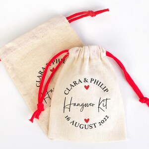 May include: Two small white cotton drawstring bags with red drawstrings. The bag on the right has the text "Clara & Philip Hangover Kit 16 August 2022" printed on it with three red hearts.