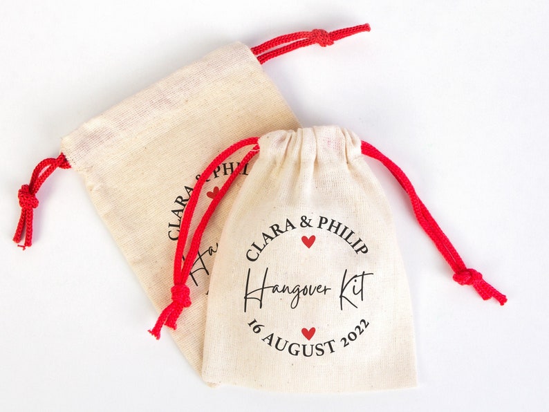 May include: Two small white cotton drawstring bags with red drawstrings. The bag on the right has the text "Clara & Philip Hangover Kit 16 August 2022" printed on it with three red hearts.