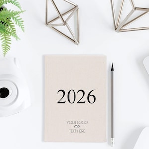 Puede incluir: Una agenda beige de 2026 con la opción de agregar un logotipo o texto, un lápiz gris, una cámara instantánea blanca y una pequeña planta en maceta. También hay formas geométricas decorativas, todo sobre una superficie blanca.
