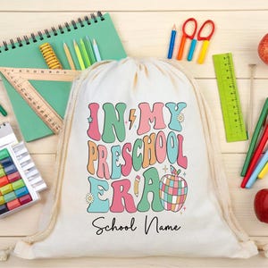 Op de afbeelding: Een witte trekkoordrugzak met de tekst "IN MY PRESCHOOL ERA" in een kleurrijk retro lettertype. De tas is omgeven door schoolspullen, waaronder potloden, kleurpotloden en appels.