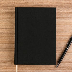 May include: A solid black rectangular notebook with a vertical spine and a single thin ribbon bookmark extending from the bottom edge, accompanied by a black ballpoint pen with a silver clip and tip positioned to the right of the notebook.