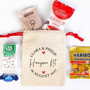 May include: A white canvas bag with red drawstring, personalized with the names Clara & Philip and the text "Hangover Kit 16 August 2022". The bag contains various items, including a small bag of Tic Tac mints, a packet of tea, a small bag of gummy candies, and a small packet of breath mints.