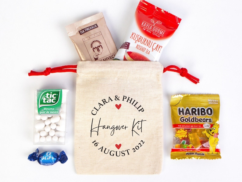 May include: A white canvas bag with red drawstring, personalized with the names Clara & Philip and the text "Hangover Kit 16 August 2022". The bag contains various items, including a small bag of Tic Tac mints, a packet of tea, a small bag of gummy candies, and a small packet of breath mints.