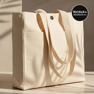 May include: A cream-colored canvas tote bag with two long shoulder straps, an exterior slip pocket, and a single dark snap button closure. The bag measures 16x14x4 inches and features a structured rectangular shape with a flat bottom base.