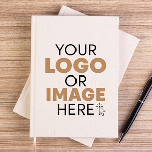 Peut inclure: Un carnet blanc sur un fond brun. Le carnet porte l'inscription "YOUR LOGO OR IMAGE HERE" imprimée sur la couverture. Un stylo noir est posé sur la table à côté du carnet.